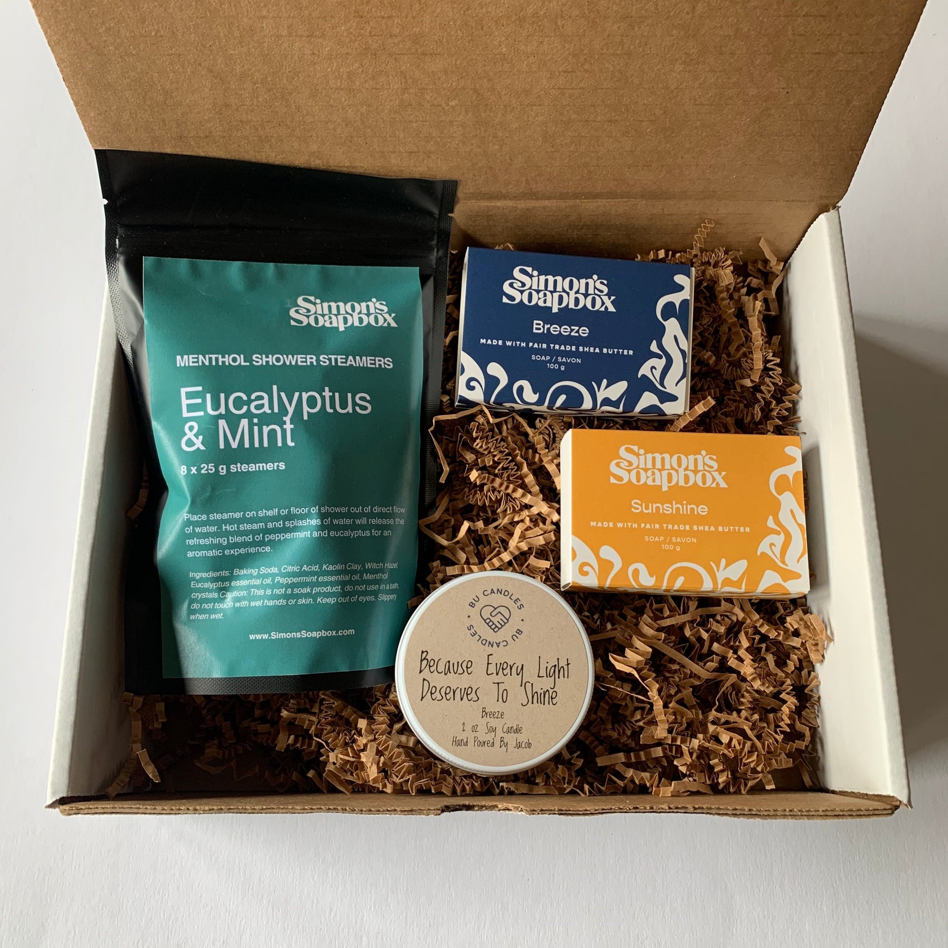 cardboard gift box with brown crinkle filled with a candle tin, black stand up pouch with green label, two bars of soap one with blue label and one with yellow label