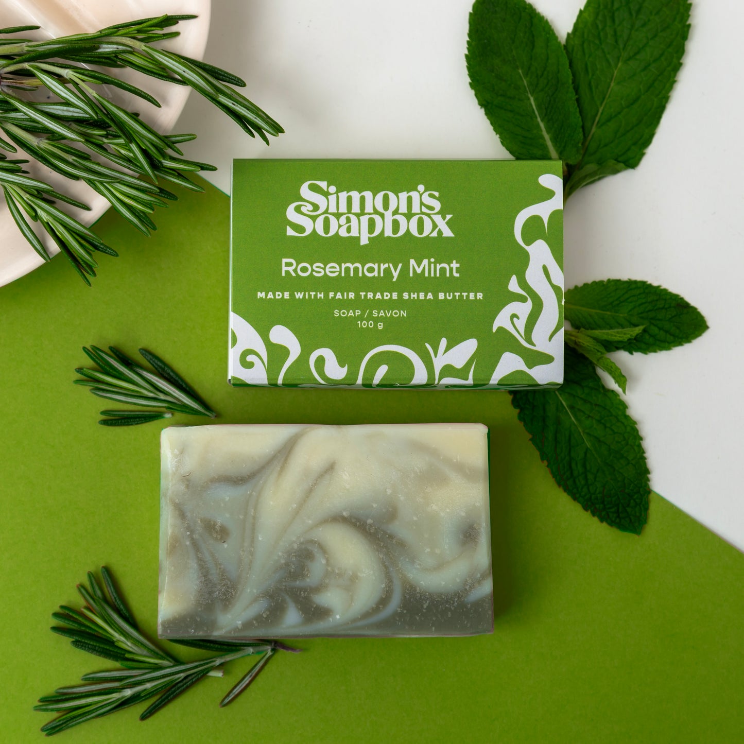 green packaged soap beside unwrapped bar of soap with green swirls. Rosemary and Mint leaves as props.
