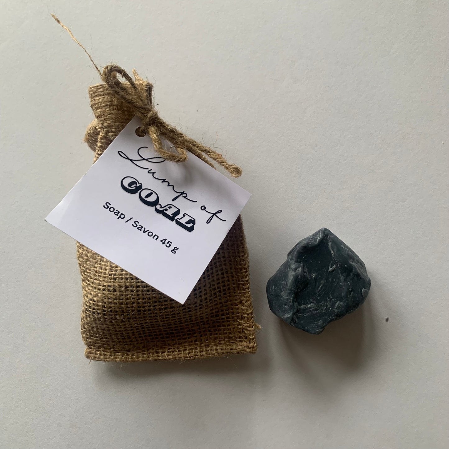 burlap drawstring bag with white label that reads "lump of coal soap" dark grey lump of soap beside bag